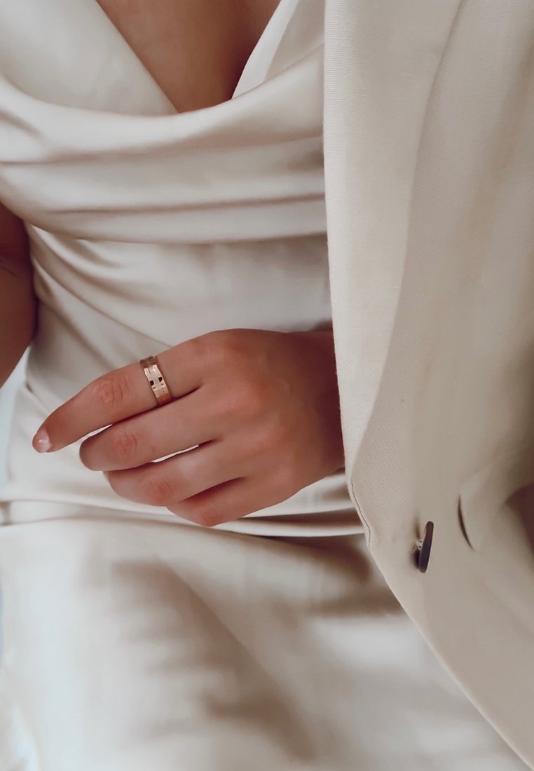 Personne en robe et veste blanches montre une bague en or à son doigt, avec un focus sur la main, la bague et les textures élégantes du tissu.