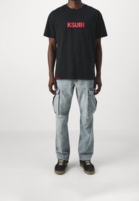 Černé bavlněné tričko s červeným logem "KSUBI". V kombinaci se světlomodrými cargo džínami se bočními kapsami a volnějším střihem. Černé tenisky.