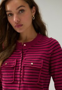 Cardigan fuchsia rayé avec des lignes noires, doté de deux poches à rabat et de boutons en métal doré, fabriqué en tissu tricot doux.