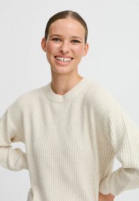 Pull en maille côtelée couleur crème avec un col rond et des manches longues, doté d'une finition texturée et de légères variations de lumière.