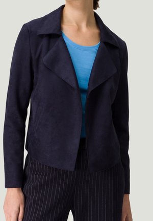Femme portant une veste ouverte bleu marine foncé, un haut bleu avec un col rond et un pantalon foncé à fines rayures sur un fond uni.
