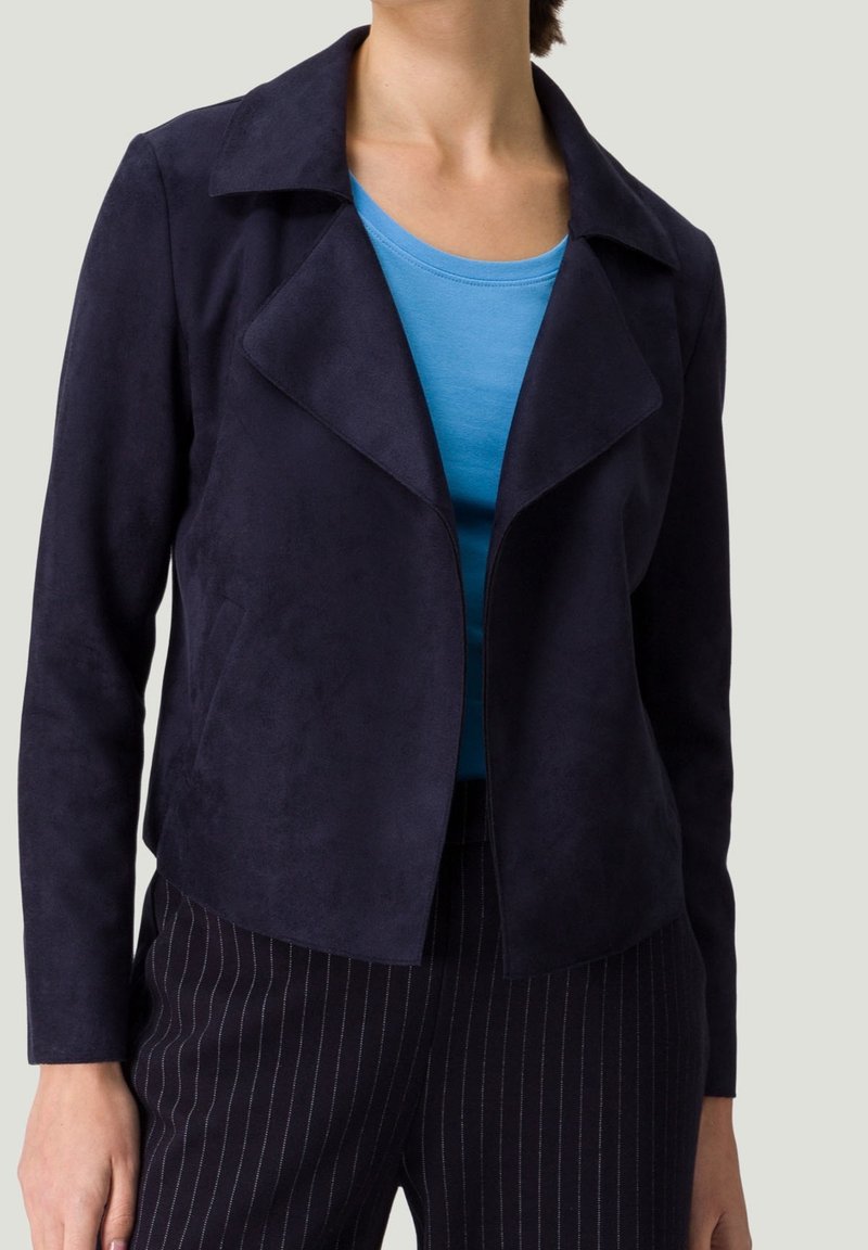 Femme portant une veste ouverte bleu marine foncé, un haut bleu avec un col rond et un pantalon foncé à fines rayures sur un fond uni.