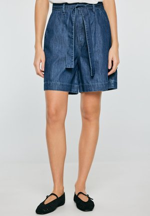 Kobieta nosząca ciemnoniebieskie dżinsowe szorty z wysokim stanem, związaną paskiem oraz czarne płaskie buty z kokardkami, stojąca na jednolitym tle.