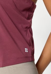 Mørkerød tanktop med en glat tekstur og forstærkede sømme, med en lille hvid logo-detalje på siden.