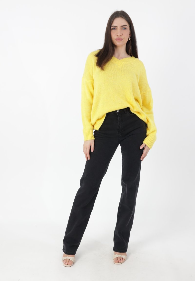 Gelber, übergroßer Strickpullover mit V-Ausschnitt und gerippten Bündchen, kombiniert mit schwarzen, geraden Jeans und beigen, riemenlosen Sandalen.