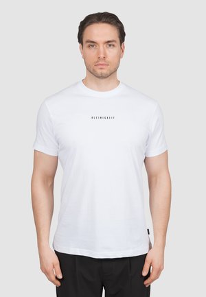 Hombre con cabello corto y castaño, vistiendo una camiseta blanca lisa con el texto "KLEINIGKEIT" centrado en el pecho y pantalones negros.