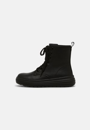 Bottine noire à lacets avec semelle épaisse à rainures et languette à l'arrière, montrée de profil sur un fond clair uni.