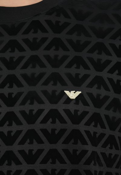 Čierne tričko s krátkym rukávom s vystupujúcim geometrickým vzorom a kovovým logom v strede. Mäkká textúra s okrúhlym výstrihom.