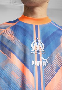 Zila un oranža sporta krekls ar teksturētu dizainu, ar augstu apkakli, "OM" logo, "DROIT AU BUT" un "PUMA" zīmolu.