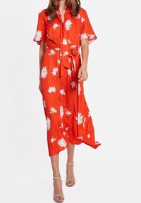 Robe rouge avec manches courtes volantées, taille ajustée par un lien, motif floral blanc. Longueur midi avec tissu fluide et texture douce.