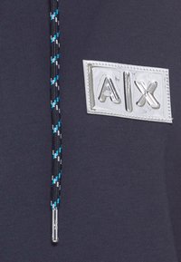 Sudadera azul marino con un parche de logo plateado que dice "A|X" en el frontal, y un cordón con un diseño trenzado en negro, blanco y azul.