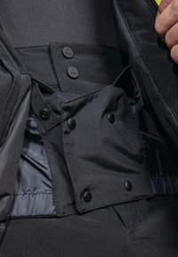 Interno di una giacca in tessuto nero che mostra bottoni a pressione, zip e una fodera liscia. Presenta una tasca con pattina e una cintura regolabile.