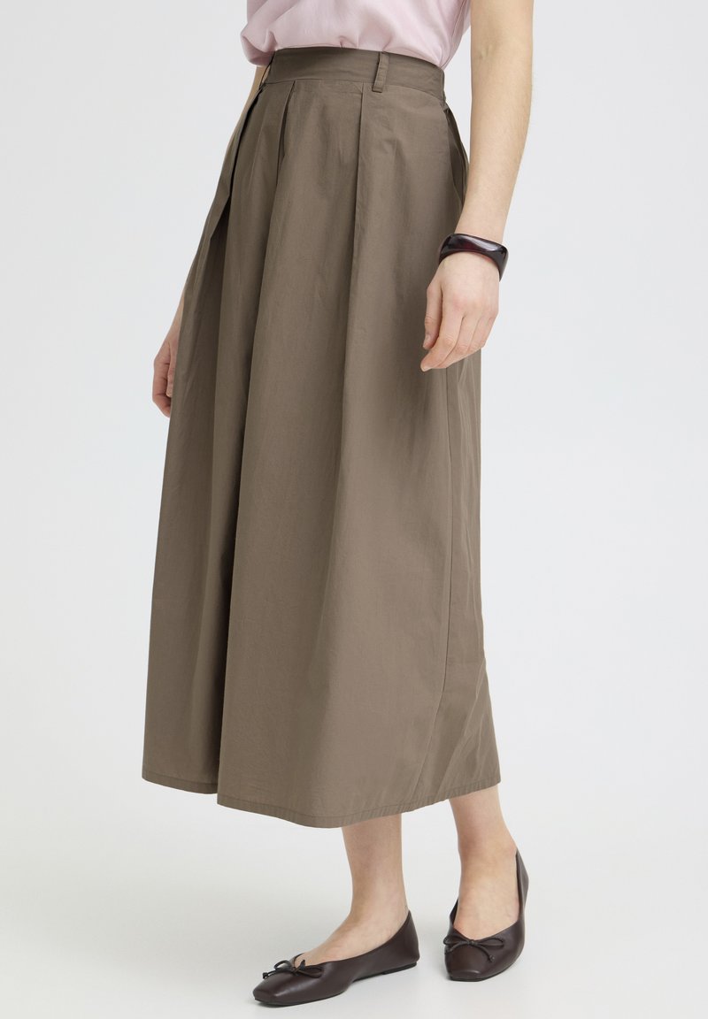 Femme portant une jupe midi taille haute verte olive avec des plis, assortie à des ballerines marron foncé et un bracelet sombre au poignet droit.