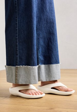 Fehér gumiszandál széles pánttal és texturált talppal, hosszú farmer szoknyával párosítva, amelynek a szegélye foszlott és kontrasztos.