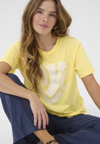 Kollane puuvillane T-särk, millel on suur valge südamekujuline "armastus" graafika. Modell istub seljas lahedad tumesinised püksid.