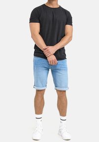 Mann trägt ein schwarzes T-Shirt, hellblaue, hochgekrempelte Jeansshorts, weiße Socken mit schwarzen Streifen und weiße Sneakers. Er steht mit gefalteten Händen.