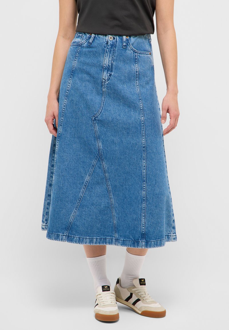 Femeie purtând o fustă din denim albastru de lungime medie, șosete albe până la gleznă, adidași bej cu dungi negre și un top negru fără mâneci, pe un fundal simplu.