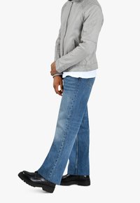 Uomo che indossa una giacca di pelle grigia, una camicia bianca, jeans a gamba ampia bicolore blu e scarpe stringate nere, in piedi di lato con le braccia incrociate.