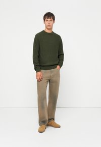Uomo che indossa un maglione lavorato a maglia verde oliva, pantaloni beige a gamba larga e sneakers color cammello, in piedi con una mano in tasca su uno sfondo bianco.