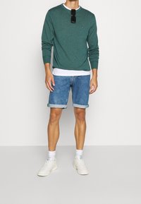 Camicia verde a maniche lunghe, maglietta bianca a collo tondo sotto, pantaloni shorts di jeans con risvolti, e sneakers bianche. Occhiali da sole sulla camicia.