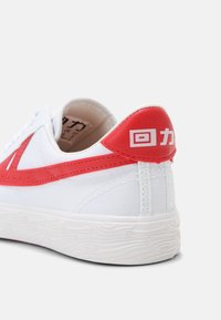 Baskets en toile blanche avec accents synthétiques rouges, semelle en caoutchouc texturé et détail du logo sur le talon. Design à lacets pour un maintien sécurisé.