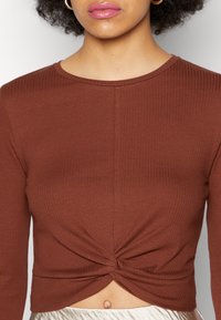 Brun ribbet langermet topp med kort lengde som har vridd frontdetalj og rund hals, og fremhever en teksturert og tettsittende design.