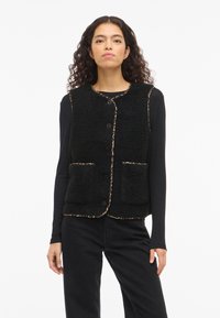 Black fleece vest featuring leopard print trim, round neckline, four buttons, and two front pockets. Worn over a long-sleeve black top.