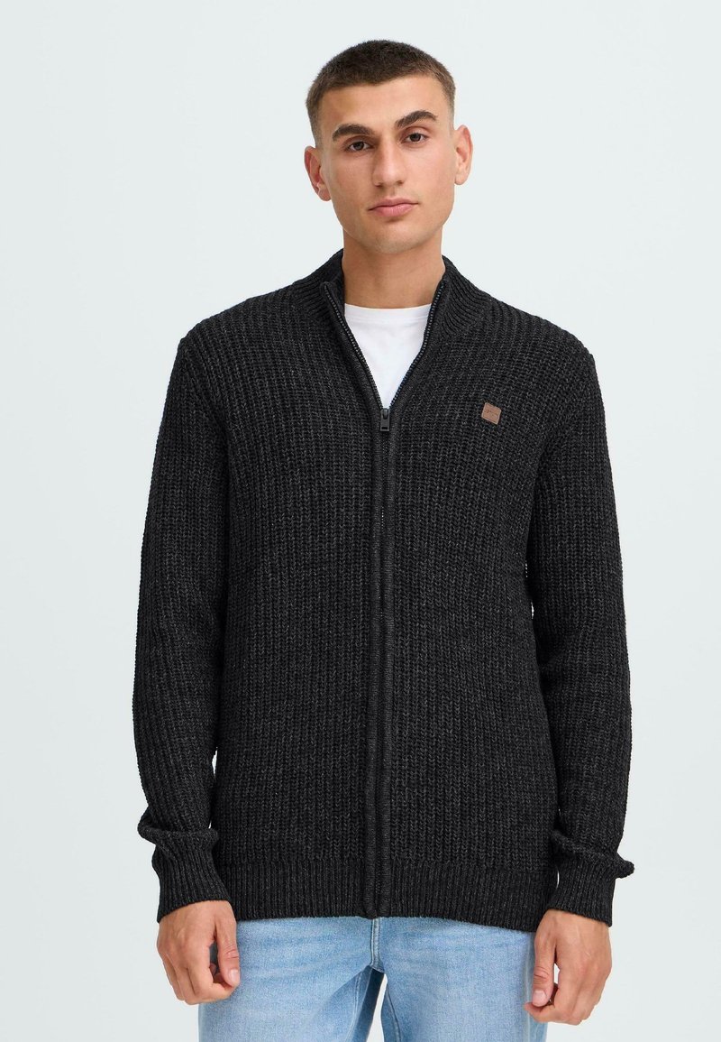 Cardigan nero in maglia con chiusura a zip, texture a coste, colletto alto e piccolo dettaglio con logo sul petto, indossato sopra una camicia bianca.