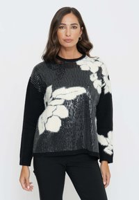 Maglione nero con un design floreale in paillettes bianche, tessuto morbido e texture, e scollo rotondo. Maniche lunghe e vestibilità rilassata.