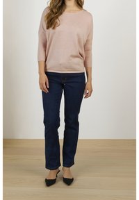 Ljusrosa tröja med trekvartsärmar, kombinerad med mörkblå jeans och svarta spetsiga skor på ett ljust trägolv.