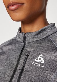 Grå dragkedja jacka med hög krage, med ett strukturerat tyg och en vit Odlo-logotyp på vänster sida.