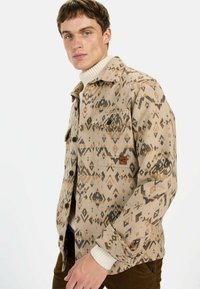 Beige gemusterde jas met een ruitmotief, voorzien van knopen, twee borstzakken en een bruine logo patch, lagen over een licht sweater.