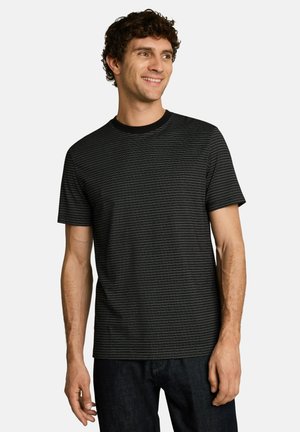 Junger Mann mit lockigem Haar, der ein schwarzes T-Shirt mit kurzen Ärmeln und dünnen horizontalen weißen Streifen trägt, lächelt und schaut nach rechts.