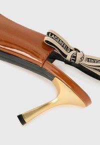 Escarpin en cuir verni marron avec un talon aiguille doré et une bride de cheville noire et beige avec un détail en nœud.