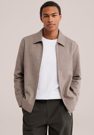 Jeune homme aux cheveux courts et bouclés portant une veste beige à fermeture éclair par-dessus une chemise blanche et un pantalon sombre, debout les mains dans les poches.