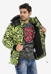 Puffjacke mit auffälligem schwarzen und neongrünen Textmuster, verfügt über einen Reißverschluss, einen verstellbaren Kordelzug am Saum und ein schwarzes Innenfutter.