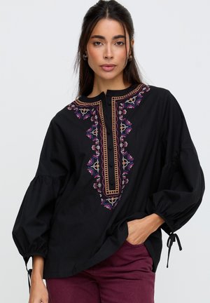 Frau trägt eine schwarze Bluse mit bunten, bestickten Mustern auf der Vorderseite und voluminösen Ärmeln, kombiniert mit dunkelroten Hosen.