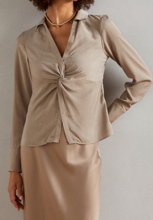 Femme portant un chemisier beige à manches longues avec un détail torsadé sur le devant et une jupe beige assortie, accessoirisée d’un collier chaîne en or.