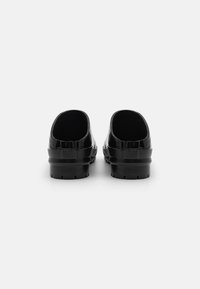 Black, glossy slip-on shoes featuring a textured band, rounded shape, and flat rubber sole with grooves. Open back design for easy wear.