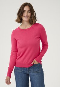 Pull à manches longues rose avec un col arrondi, des poignets et un ourlet côtelés. Porté avec un jean bleu, mettant en avant une coupe décontractée. Texture douce.