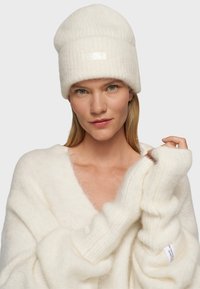 Bonnet côtelé blanc avec un revers et un patch logo. Le modèle porte un pull en blanc, doux et surdimensionné, avec des manches larges.