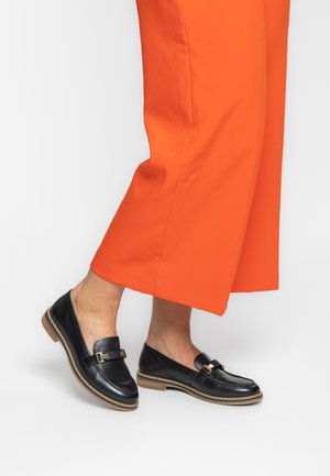 Black leather loafers with gold hardware accents, featuring a rounded toe and flat sole. Worn with bright orange wide-leg pants.
