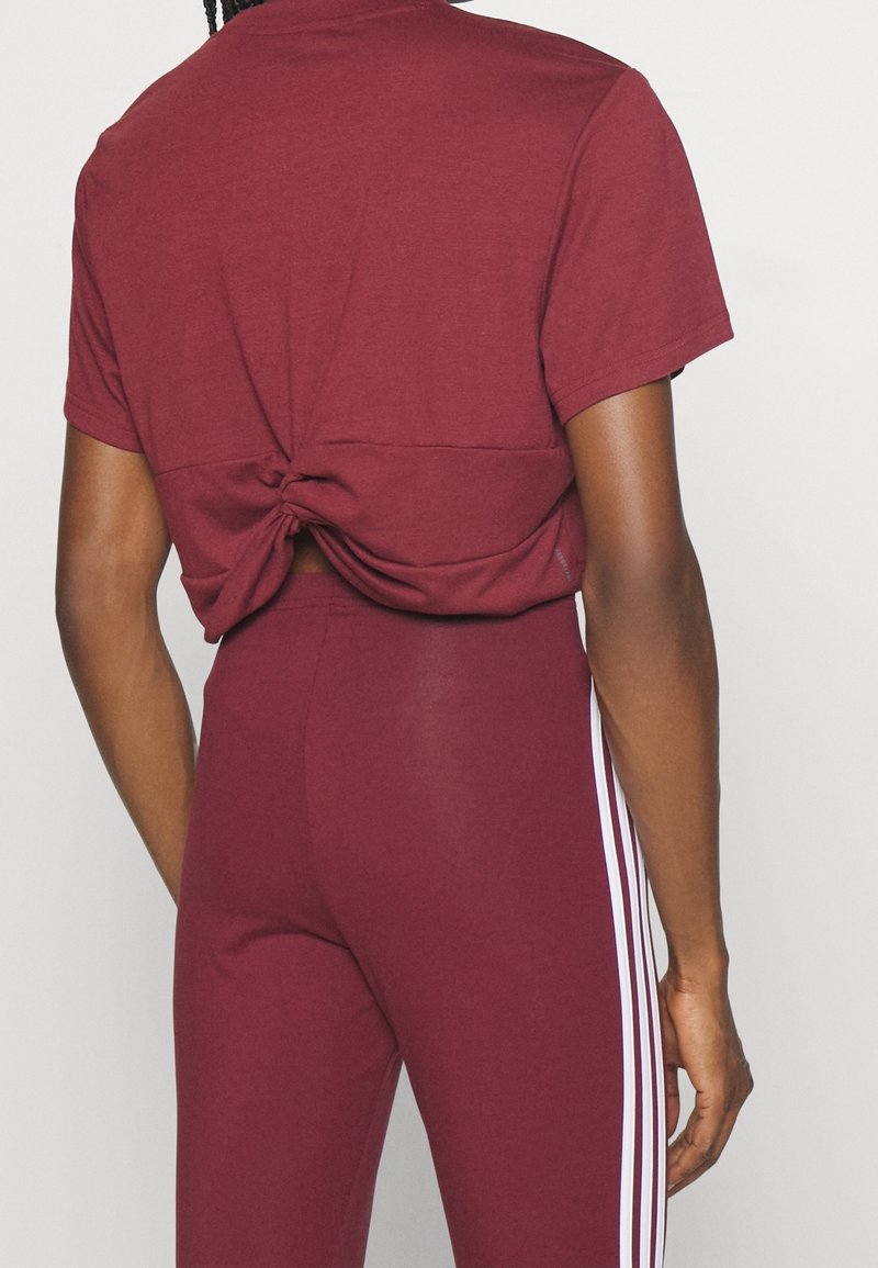 Personne portant un crop top bordeaux noué et un legging taille haute assorti avec des bandes blanches sur les côtés, vue de dos.