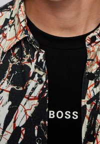 Ein Hemd mit Knöpfen, das mit einem schwarz-weißen und roten abstrakten Muster versehen ist, über einem schwarzen T-Shirt getragen wird, das in fettem Weiß den Text "BOSS" zeigt.