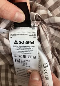 Braun-weißer kariert Stoff mit Pflegeetikett, das den Markennamen "Schöffel" und die Waschanweisungen zeigt. Das Etikett ist am Kragen befestigt.