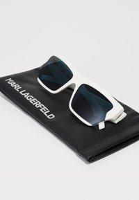 Valged ristkülikukujulised prillid tumedate läätsedega, asetatud mustale kaitsekotile, millel on valgete tähtedega tekst "KARL LAGERFELD".