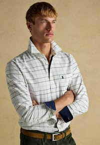 Weiß kariertes Hemd mit blauen Linien, ausgestattet mit einem Button-Down-Kragen und kontrastierenden marineblauen Manschetten, kombiniert mit einem braunen Gürtel und grüner Hose.