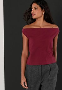 Burgundijas krāsas topiņš ar atvērtām plecu daļām, izgatavots no pieguļoša kokvilnas materiāla, ar īsām piedurknēm un gludu tekstūru, valkāts kopā ar pelēkām piegriezuma biksēm.