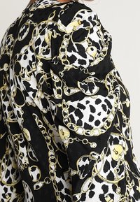 Blouse noir et blanc avec imprimé léopard et motif chaîne dorée, dotée de manches bouffantes et d'un tissu à texture lisse.