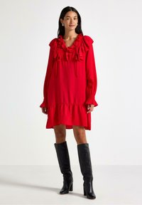 Rød langærmet kjole med flæser ved halsudsk�æringen og bunden, der har en løstsiddende pasform og en struktureret stof. Bæres med knæhøje sorte støvler.